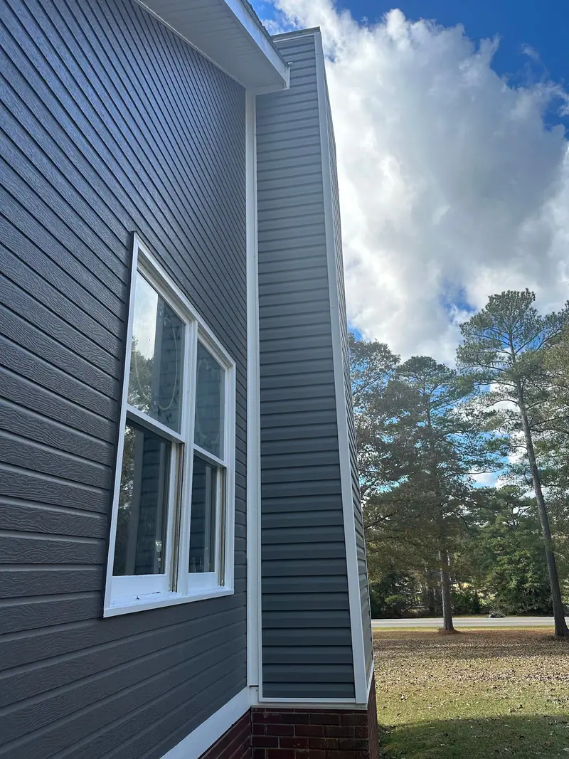 Tall wall of dark gray lap siding with a white-trimmed window and pine trees behind, a Mosqueda Construction siding job