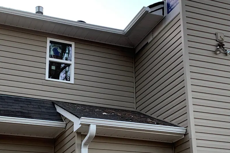 New window in tan lap siding beside a white gutter and downspout, a Mosqueda Construction job
