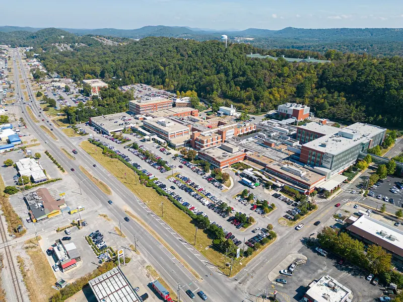 A sunny drone view along a divided multi-lane highway lined with shops and a Shell station, with a large brick hospital campus and full car parks in the centre and wooded hills and a water tower behind.