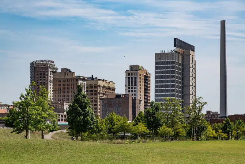 Downtown high-rises (one with a rooftop "CITY FEDERAL" sign) and a tall slim smokestack seen across a green park lawn and young trees under a bright, lightly clouded sky.