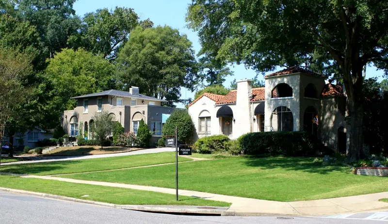 Two stucco houses, one with a red clay-tile roof and arched tower, on a sunny tree-shaded residential street corner with clipped lawns; the corner street sign reads "Bonita Drive / Poinciana Drive".
